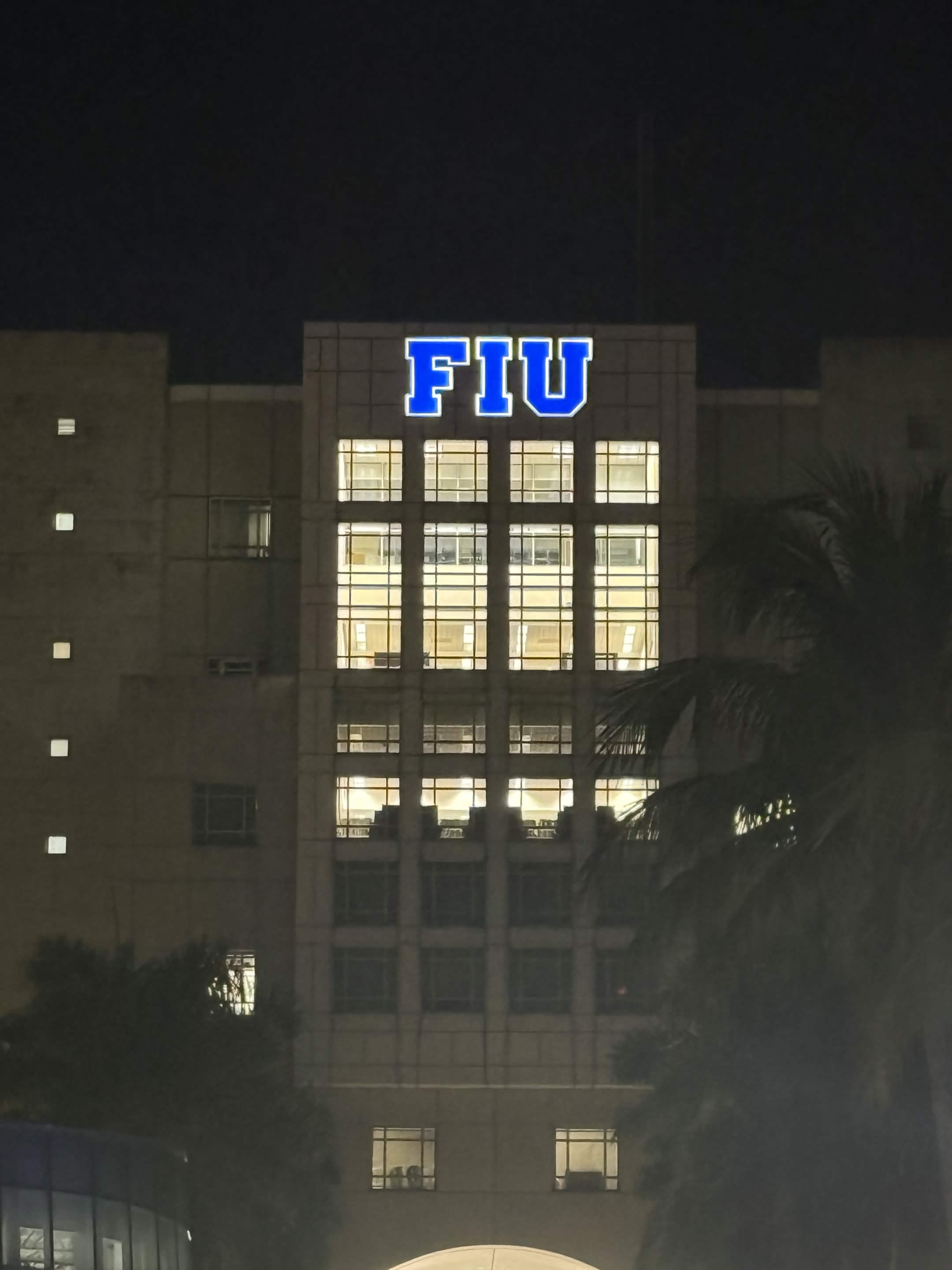 The FIU sign lit up blue on a campus building at night